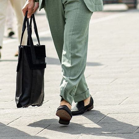 male model holding tote bag, legs walking on street wearing trousers and loafers