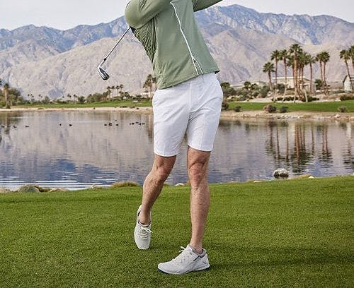 Male golfer swinging a golf club wearing a zip up sweater, shorts and baseball cap