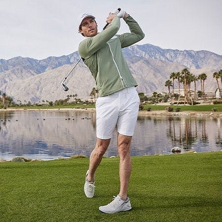 Male golfer swinging a golf club wearing a zip up sweater, shorts and baseball cap