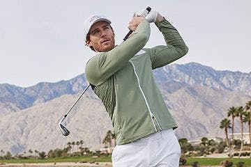 Male golfer swinging golf club wearing a zip up sweater, shorts and baseball cap