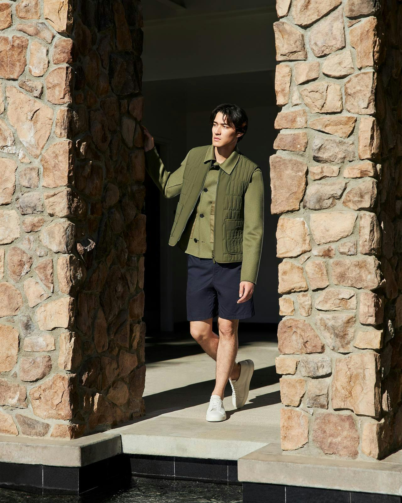 a man in a jacket and shorts standing in front of a stone wall