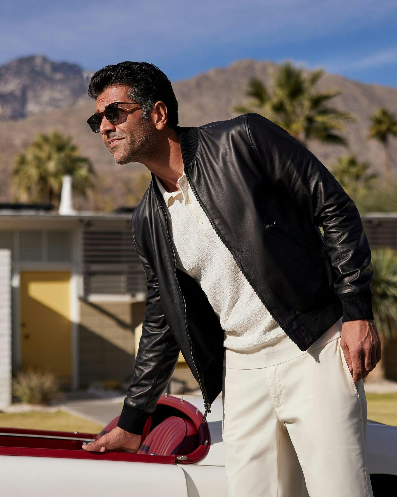 a man wearing a white outfit and leather jacket  leaning on a car